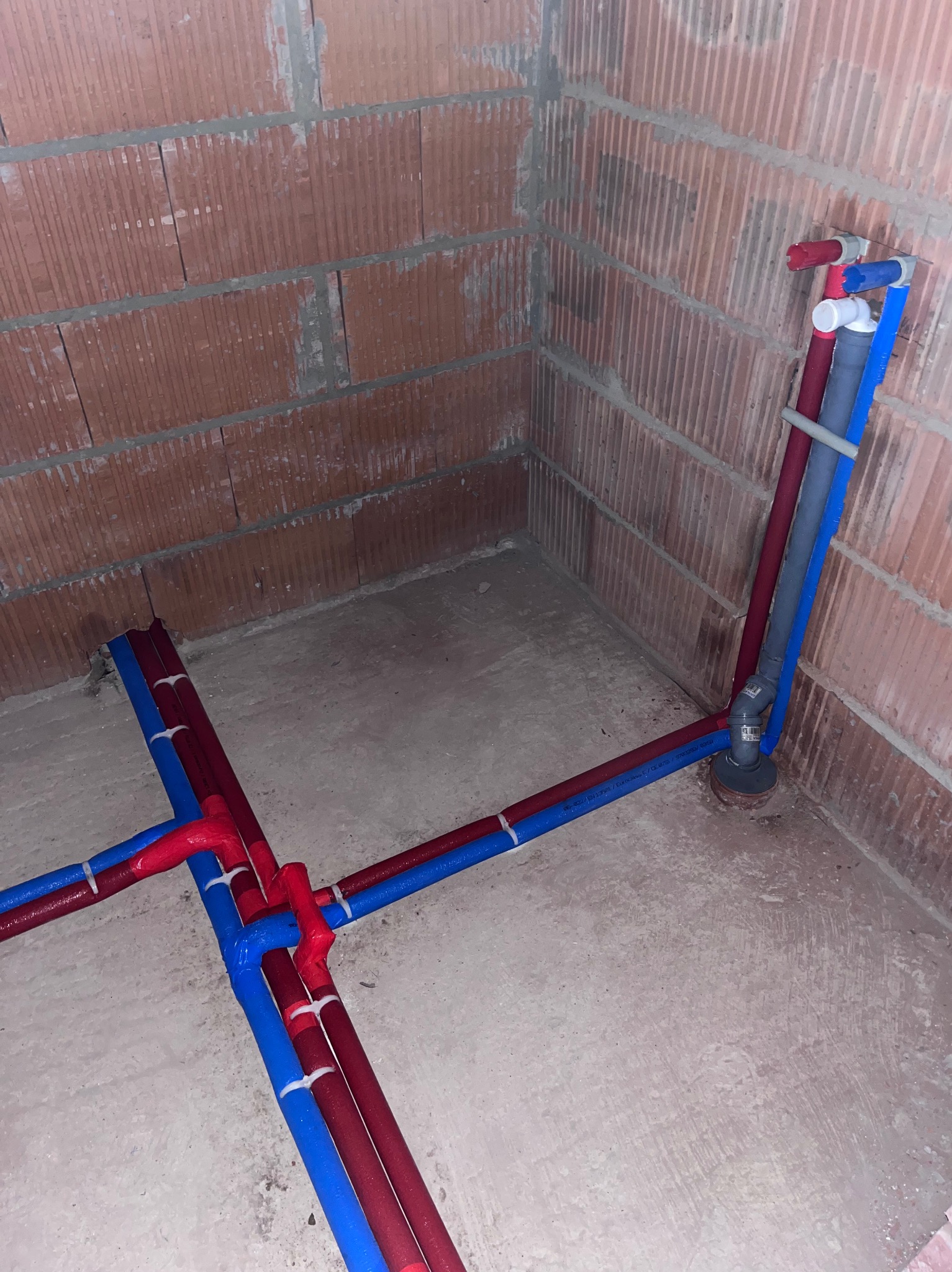 Surowe wnętrze z widocznymi, częściowo zamontowanymi rurami instalacji wodnej i grzewczej, z czerwonymi i niebieskimi rurami wychodzącymi ze ściany i podłogi, w tle ściana z cegły dziurawki.