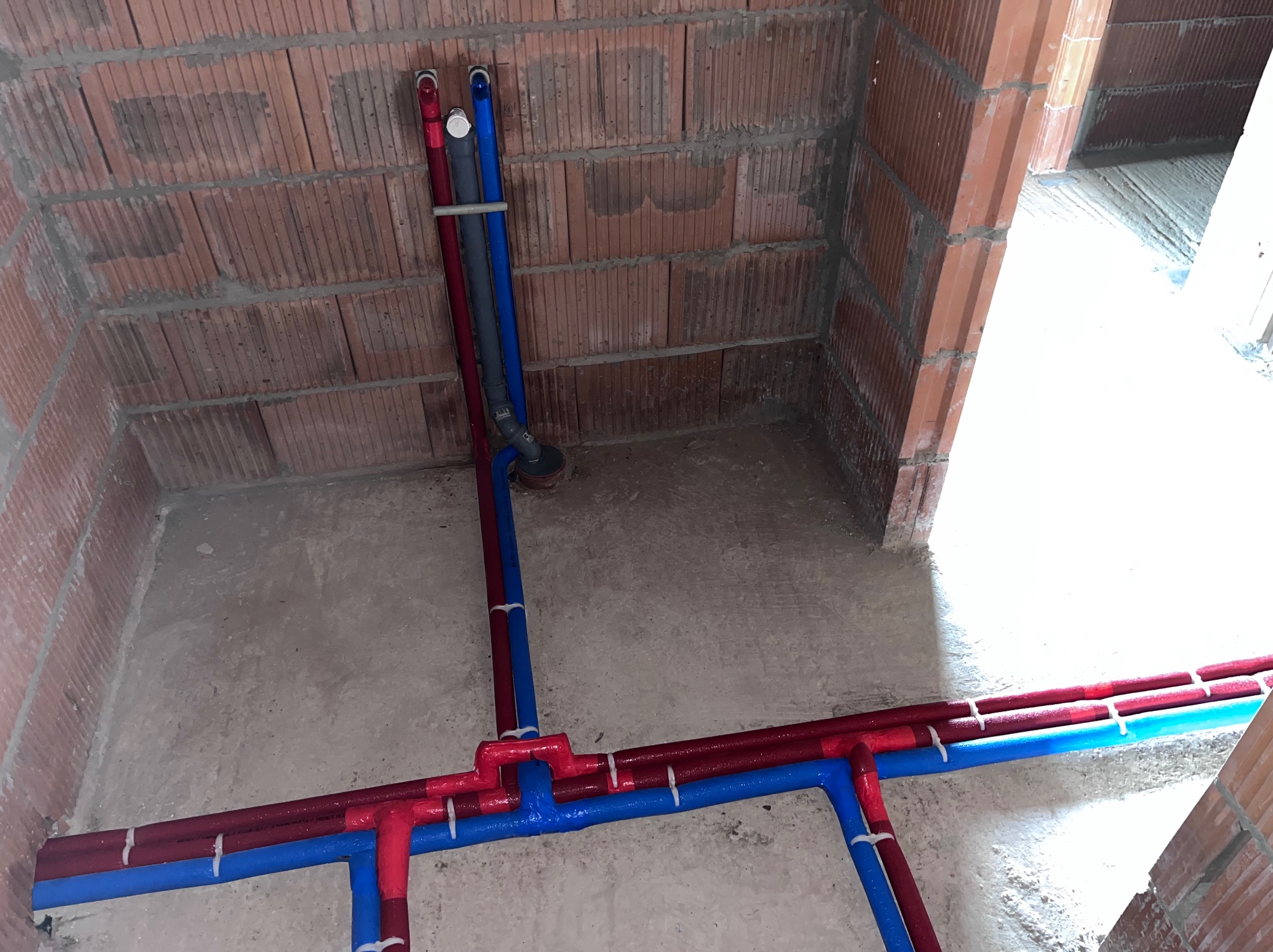 Surowy stan instalacji hydraulicznej w nowo budowanym domu, widoczne rury doprowadzające wodę w kolorach czerwonym i niebieskim, przymocowane do ściany z cegły kratówki za pomocą białych opasek...