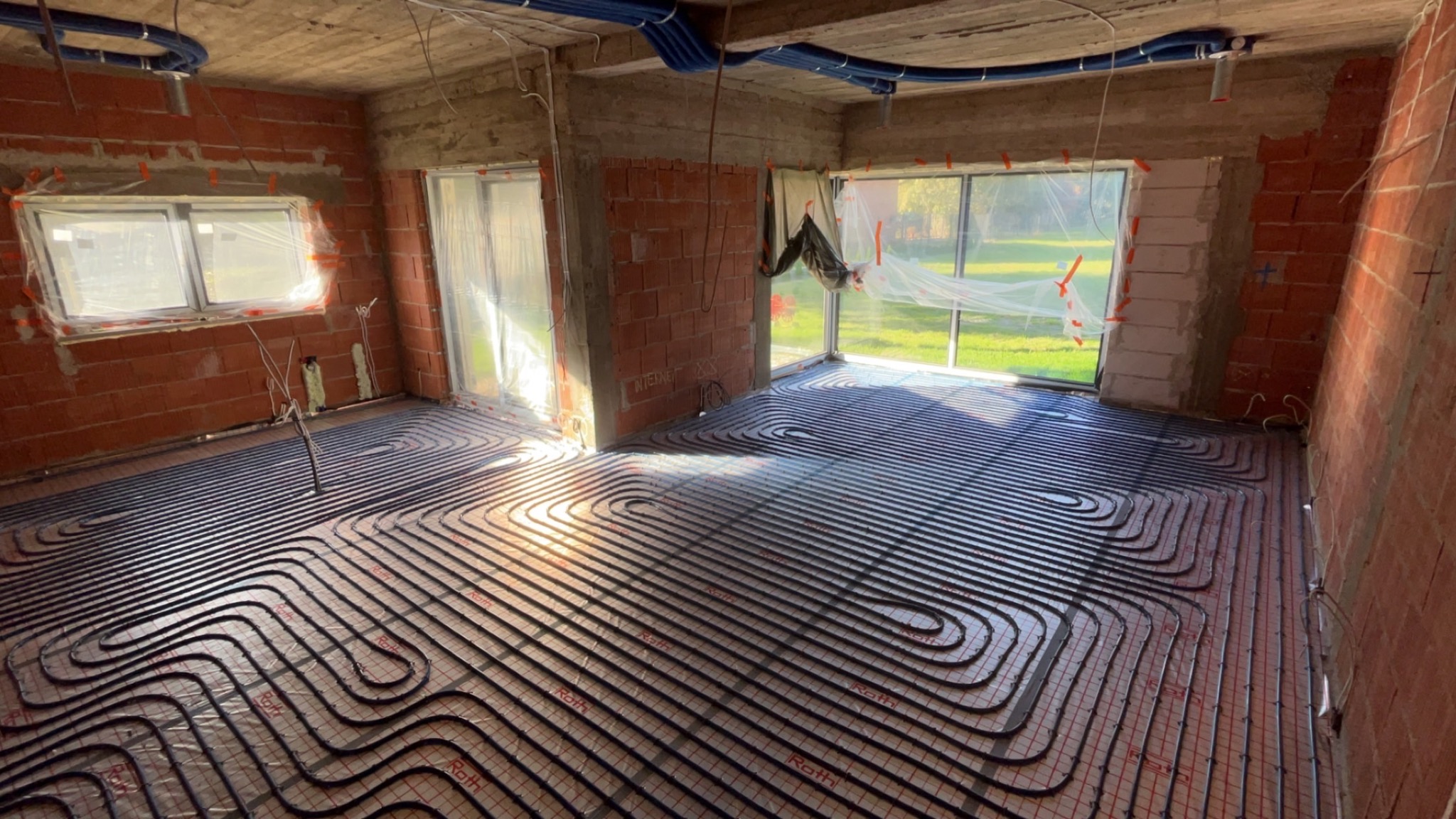 Wnętrze w stanie surowym z rozłożoną instalacją ogrzewania podłogowego Roth na folii z siatką, widoczne okna zabezpieczone folią, ściany z czerwonej cegły.