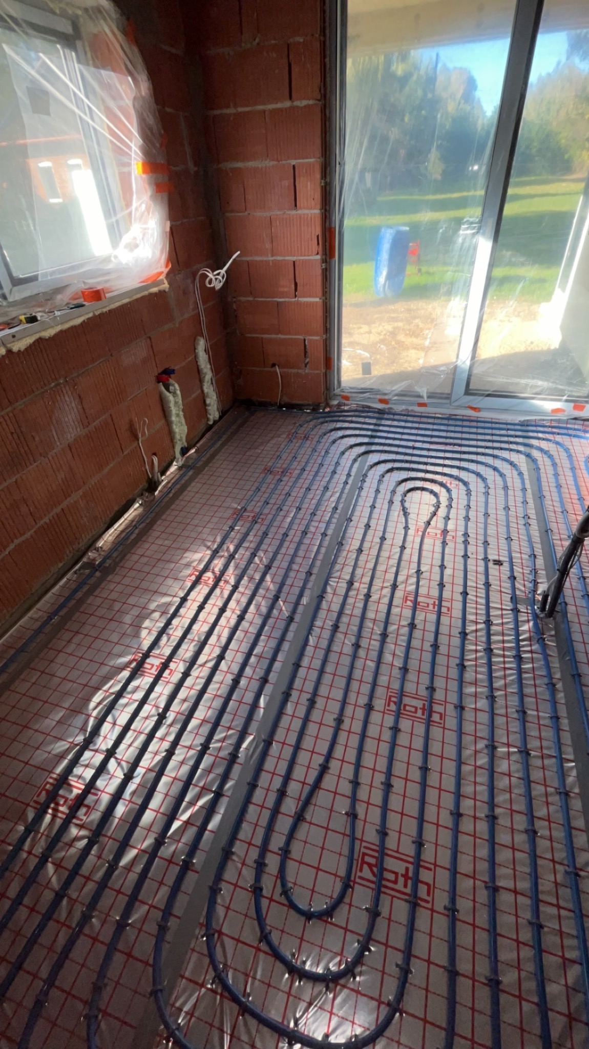 Widok na podłogę z rozprowadzoną instalacją ogrzewania podłogowego z rurami Roth, na podkładzie z siatką w nowo budowanym pomieszczeniu z ceglanymi ścianami i dużym oknem.
