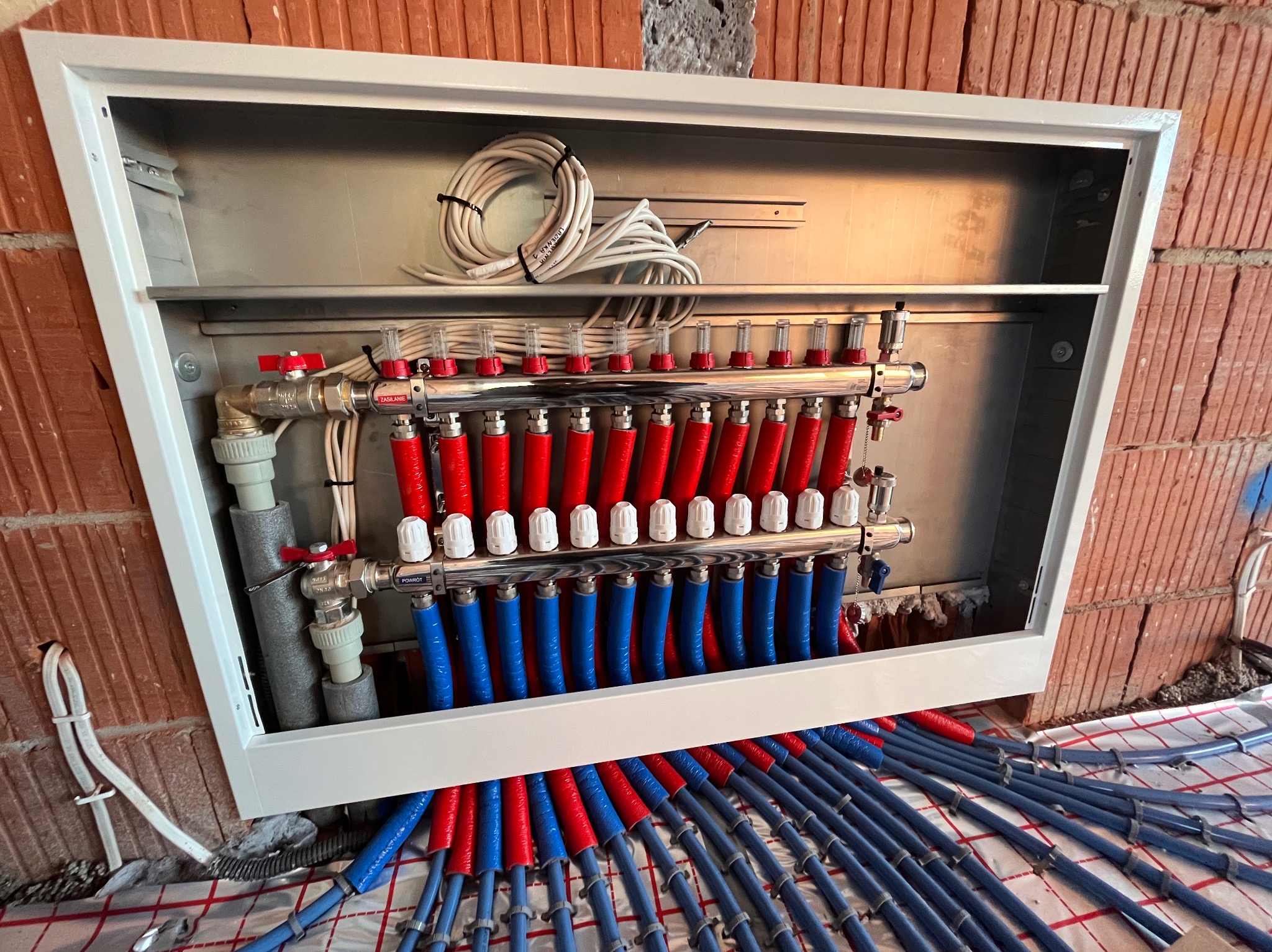 Rozdzielacz ogrzewania podłogowego w białej skrzynce, z widocznymi czerwonymi i niebieskimi rurami, zamontowany na ścianie z cegły, z przewodami elektrycznymi na górze.