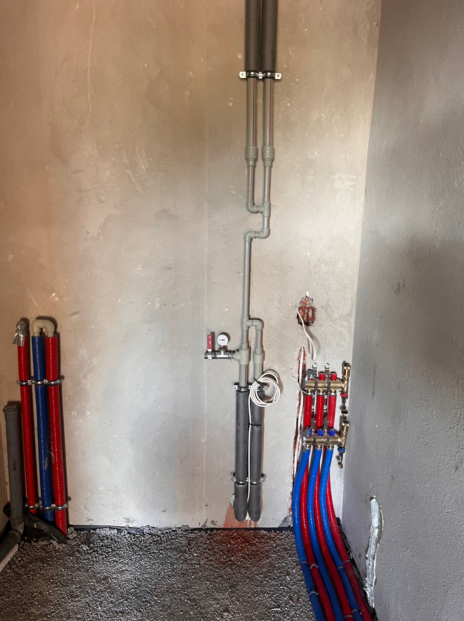 Wnętrze w trakcie instalacji ogrzewania podłogowego: widoczne rury grzewcze w kolorze czerwonym i niebieskim, rozdzielacz mosiężny, szare rury pionowe przy ścianie i wylewka podłogowa z kamyczków.