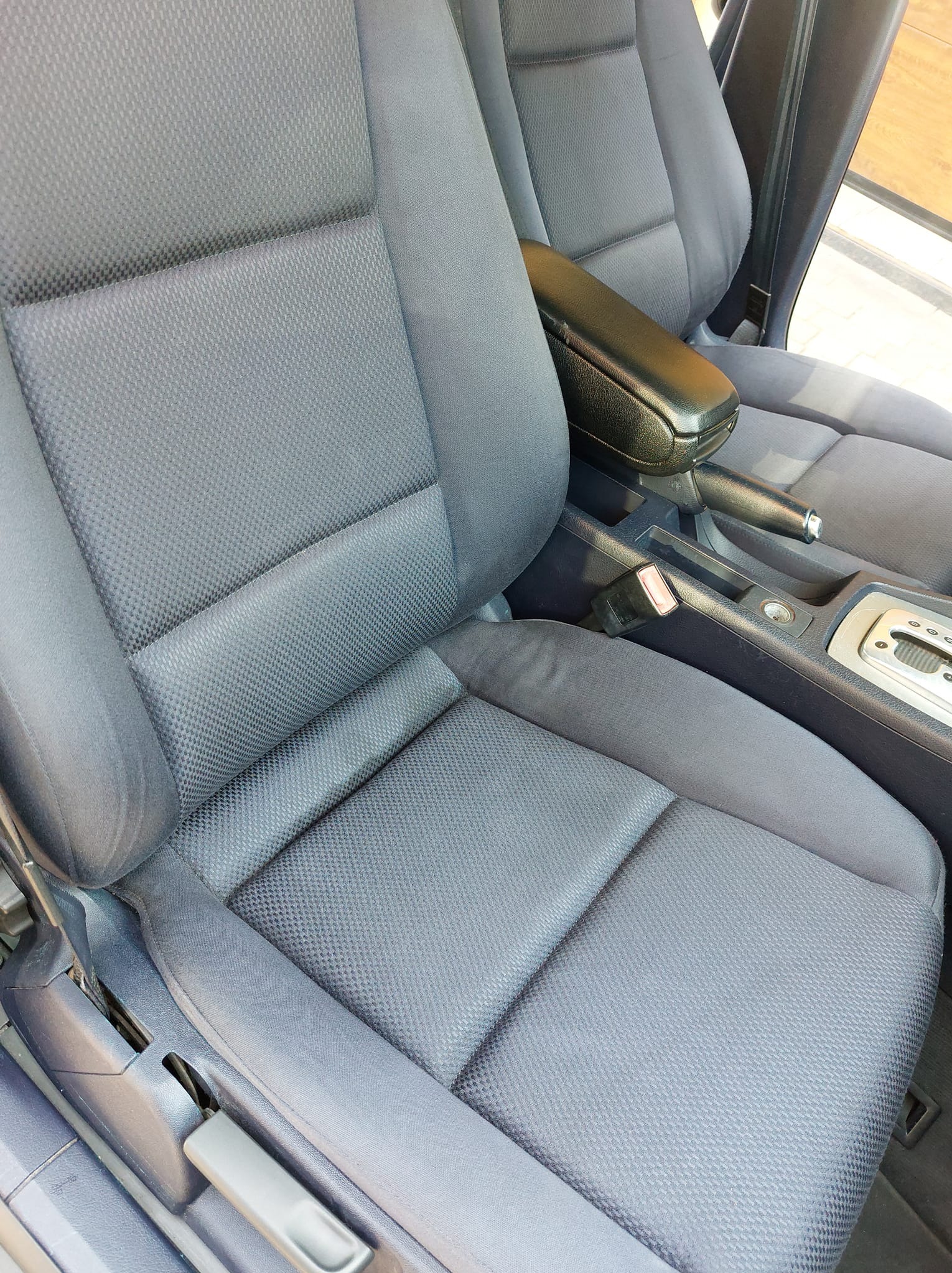 Szare, tekstylne siedzenie samochodowe z czarnym podłokietnikiem, widoczny mechanizm regulacji, pas bezpieczeństwa i dźwignia hamulca ręcznego.
