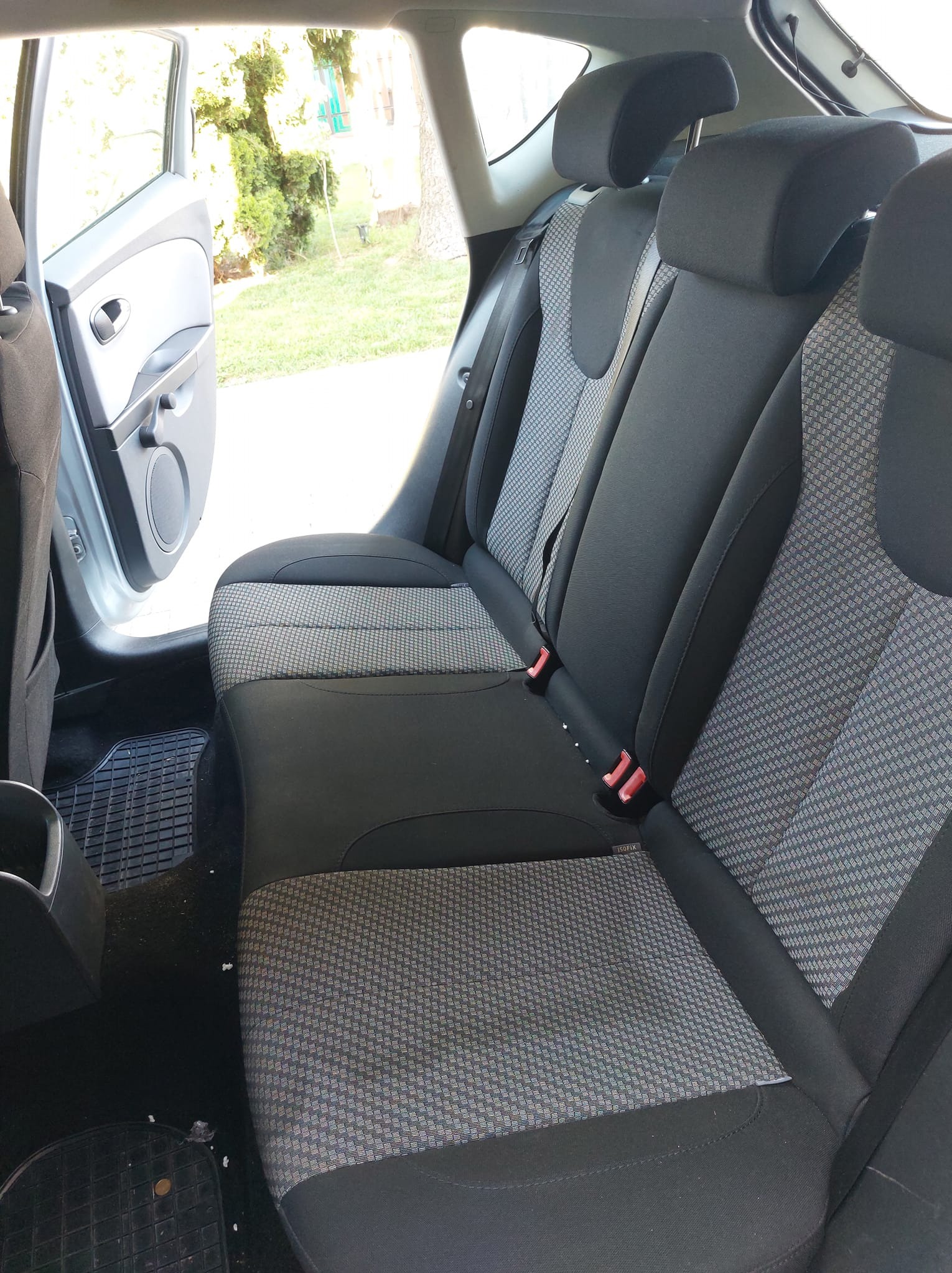 Wnętrze samochodu z widocznymi tylnymi siedzeniami z tapicerką w szaro-czarną kratę i otwartymi drzwiami po lewej stronie, przez które widać zieleń drzew.