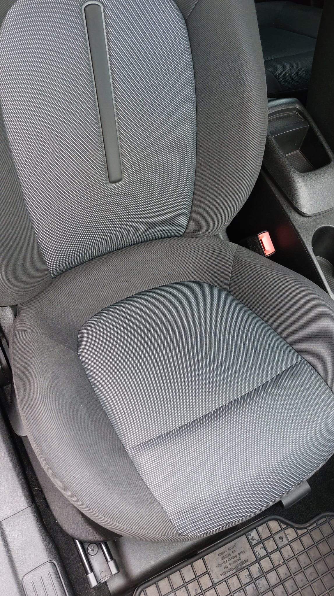 Szare, tekstylne siedzenie samochodowe z widocznym zagłówkiem, pasem bezpieczeństwa i gumową wycieraczką pod nogami.