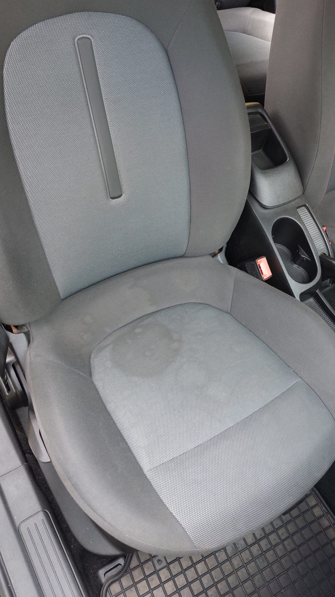 Szare, tekstylne siedzenie samochodowe z widocznymi plamami przed czyszczeniem, zapięcie pasa bezpieczeństwa w tle.