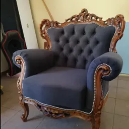 Elegancki fotel w stylu Ludwika z ciemnoszarą tapicerką i rzeźbioną, drewnianą ramą, wykończony srebrnymi pinezkami, na tle żółtej ściany i podłogi z płytek.