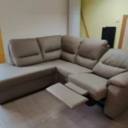 Beżowy narożnik z wysuwanym podnóżkiem, ustawiony w pomieszczeniu z beżowymi i żółtymi ścianami, widoczne okno i niezidentyfikowane przedmioty w tle.