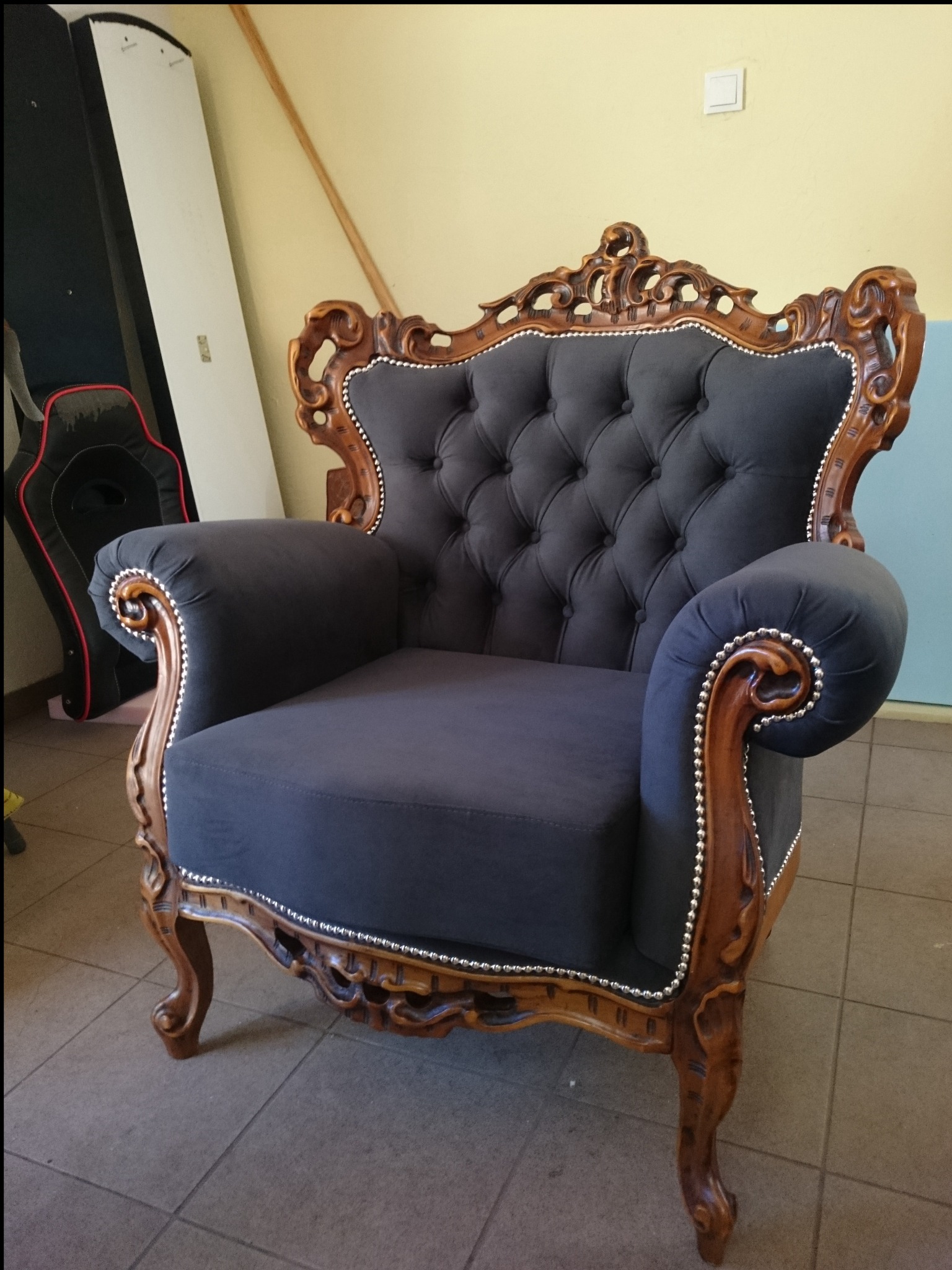 Elegancki fotel w stylu Ludwika z ciemnoszarą tapicerką i rzeźbioną, drewnianą ramą, wykończony srebrnymi pinezkami, na tle żółtej ściany i podłogi z płytek.