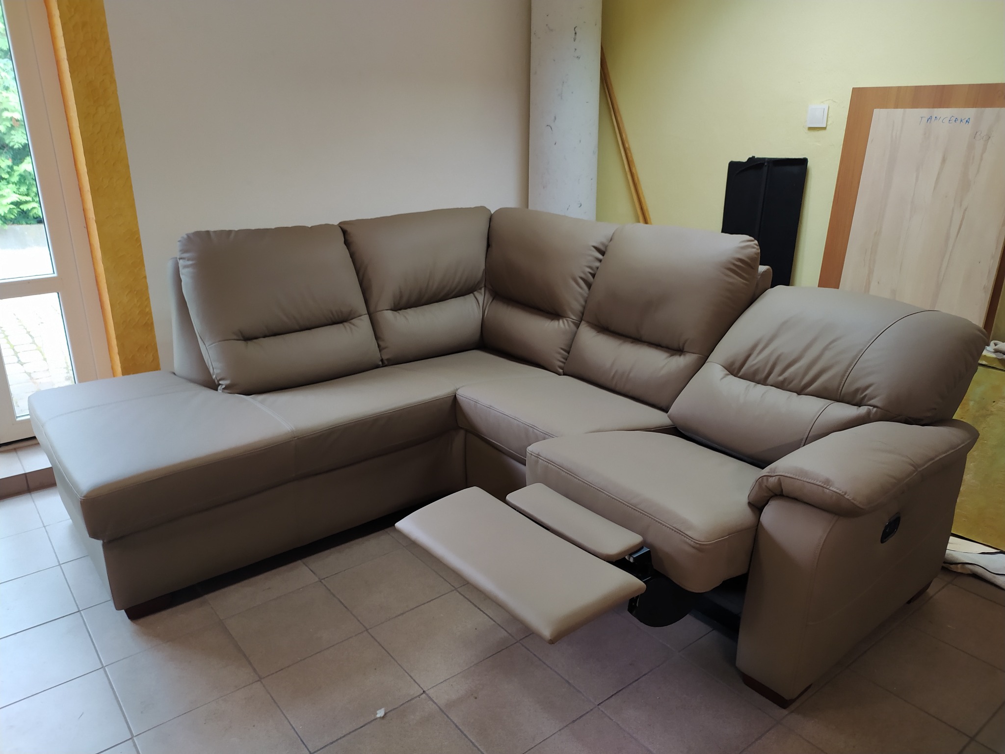 Beżowy narożnik z wysuwanym podnóżkiem, ustawiony w pomieszczeniu z beżowymi i żółtymi ścianami, widoczne okno i niezidentyfikowane przedmioty w tle.