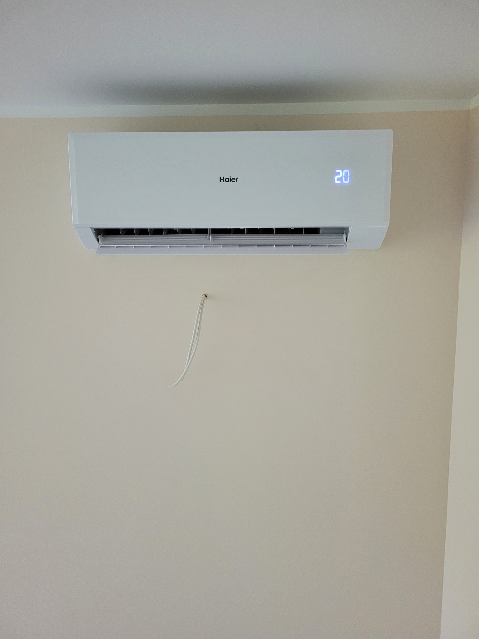 Biały klimatyzator Haier na beżowej ścianie w mieszkaniu, widoczne przewody. Wyświetlacz pokazuje temperaturę 20 stopni.