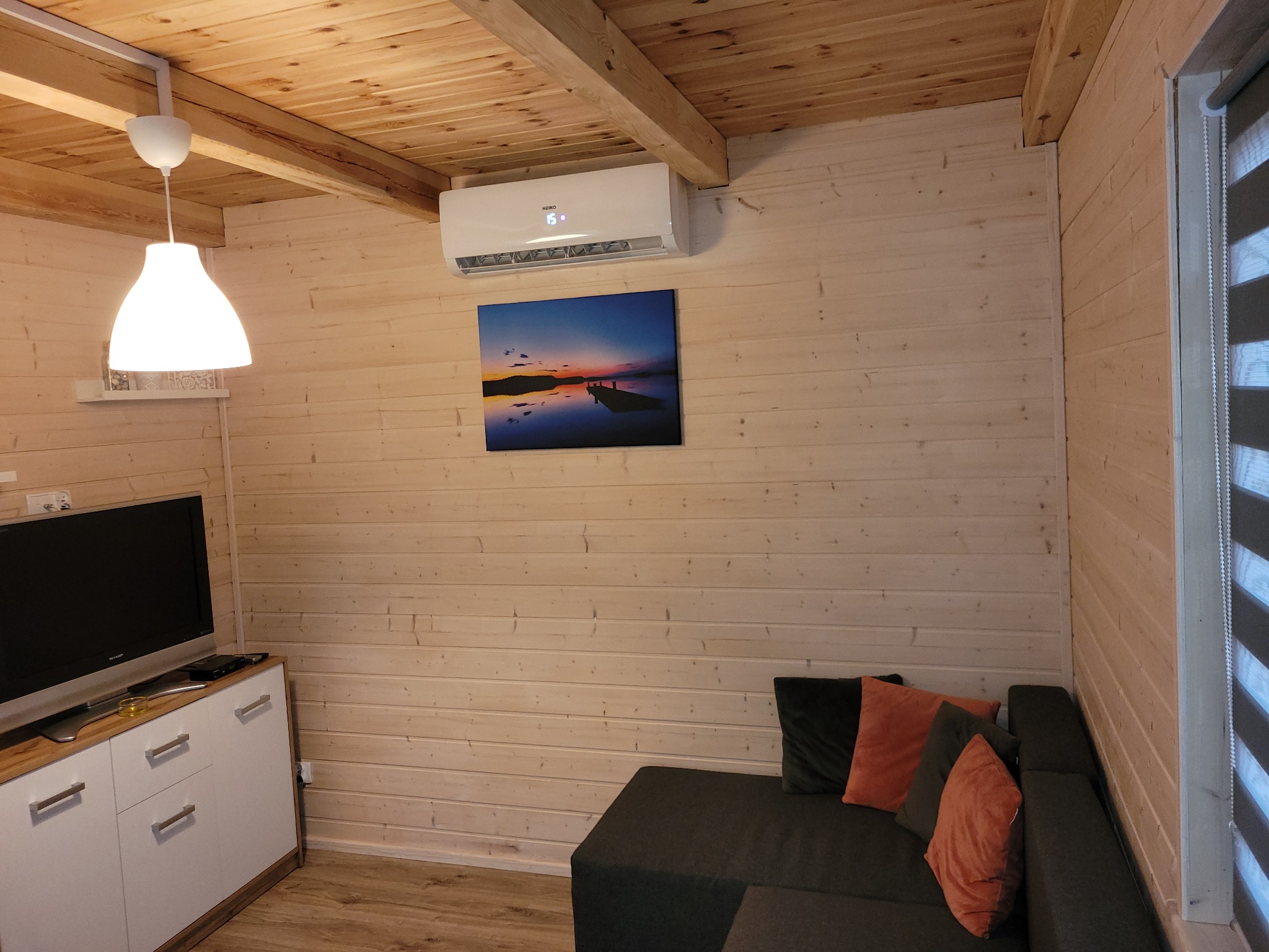 Wnętrze domku letniskowego z klimatyzatorem na ścianie z boazerii, nad sofą w kolorach szarości i pomarańczy. Widoczny telewizor i drewniany sufit.