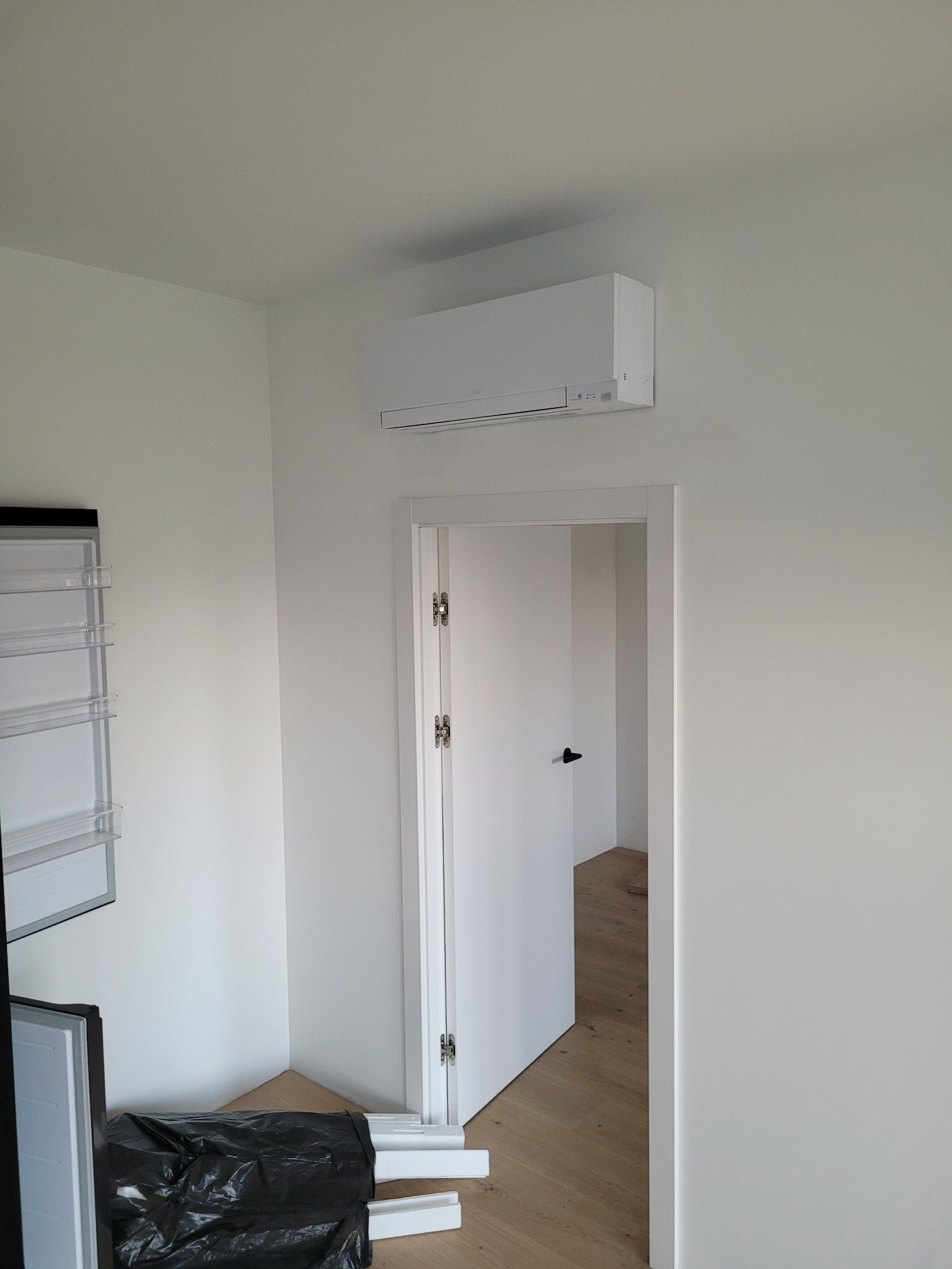 Biały klimatyzator ścienny w minimalistycznym wnętrzu z widokiem na otwarte drzwi. Częściowo widoczna lodówka i śmieci po remoncie.