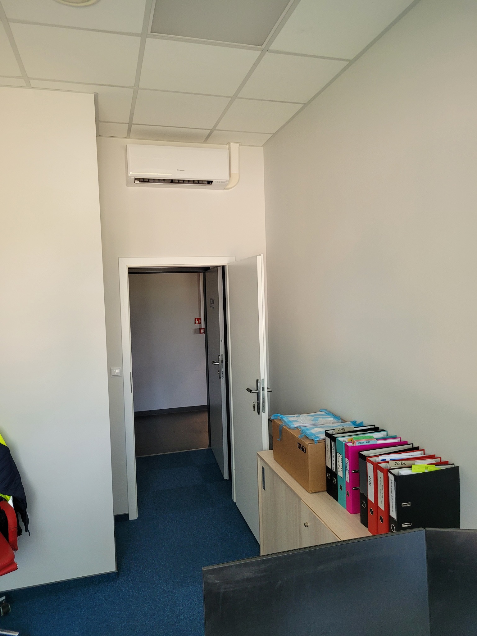 Biała klimatyzacja ścienna zamontowana w biurze nad drzwiami. Widoczne segregatory na szafce i fragment biurka z monitorem. Niebieska wykładzina podłogowa.