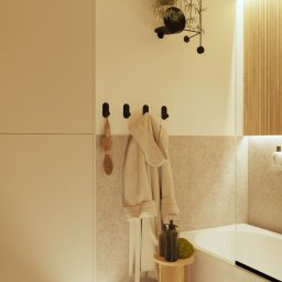 eMPe Pracownia sztuki - kreowanie przestrzeni wnętrz Paulina A. Matyja - Jasna łazienka z wanną, drewnianym akcentem na ścianie, rośliną w nowoczesnej doniczce na ścianie, beżowym ręcznikiem i stołkiem z kosmetykami, oświetlenie LED przy podłodze.