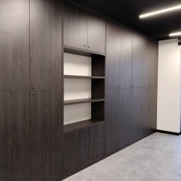 Bartłomiej Mucha - Długa ściana zabudowana ciemnymi szafami z drewnianymi frontami, z otwartą wnęką z półkami, oświetlenie sufitowe, szara podłoga