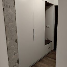 Bartłomiej Mucha - Biała szafa wnękowa z czarnymi uchwytami, widoczny fragment ściany z surowego betonu z napisami, drewnopodobna podłoga i czarny worek na śmieci w rogu pomieszczenia.