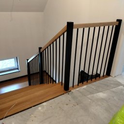 Bartłomiej Mucha - Nowoczesne schody z drewnianymi stopniami i czarną balustradą, widok z góry na dół, z oknem w tle i śladami prac wykończeniowych na podłodze.