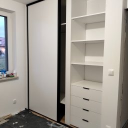 Bartłomiej Mucha - Biała szafa wnękowa z przesuwnymi drzwiami i czarnymi obramowaniami, widoczne półki i szuflady, podłoga zabezpieczona folią malarską, okno z widokiem na dom w tle.