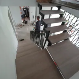 Wnętrze w trakcie wykańczania: widok z góry na schody z drewnianymi stopniami i czarną, metalową balustradą o geometrycznym wzorze, w tle widoczni dwaj pracownicy podczas prac malarskich.