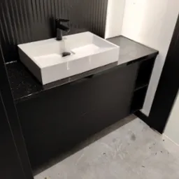 Nowoczesna, prostokątna umywalka nablatowa z czarną armaturą na czarnej szafce z blatem w czarne plamki, na tle czarnej ściany z pionowymi żłobieniami.