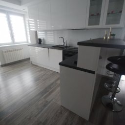 Bartłomiej Mucha - Biała, nowoczesna kuchnia z barkiem i hokerami. Fronty szafek na wysoki połysk, szare panele podłogowe imitujące drewno. Minimalistyczny design, jasne wnętrze.