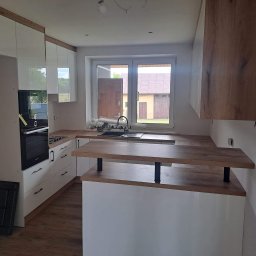 Bartłomiej Mucha - Jasna kuchnia z drewnianymi akcentami: białe szafki, drewniany blat i wyspa kuchenna z czarnymi wspornikami. Okno z widokiem na budynek gospodarczy.