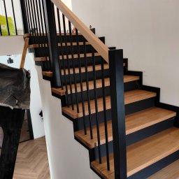Bartłomiej Mucha - Nowoczesne schody z drewnianymi stopniami i czarną balustradą, widok z dołu, parkiet w jodełkę na podłodze, minimalistyczny design.