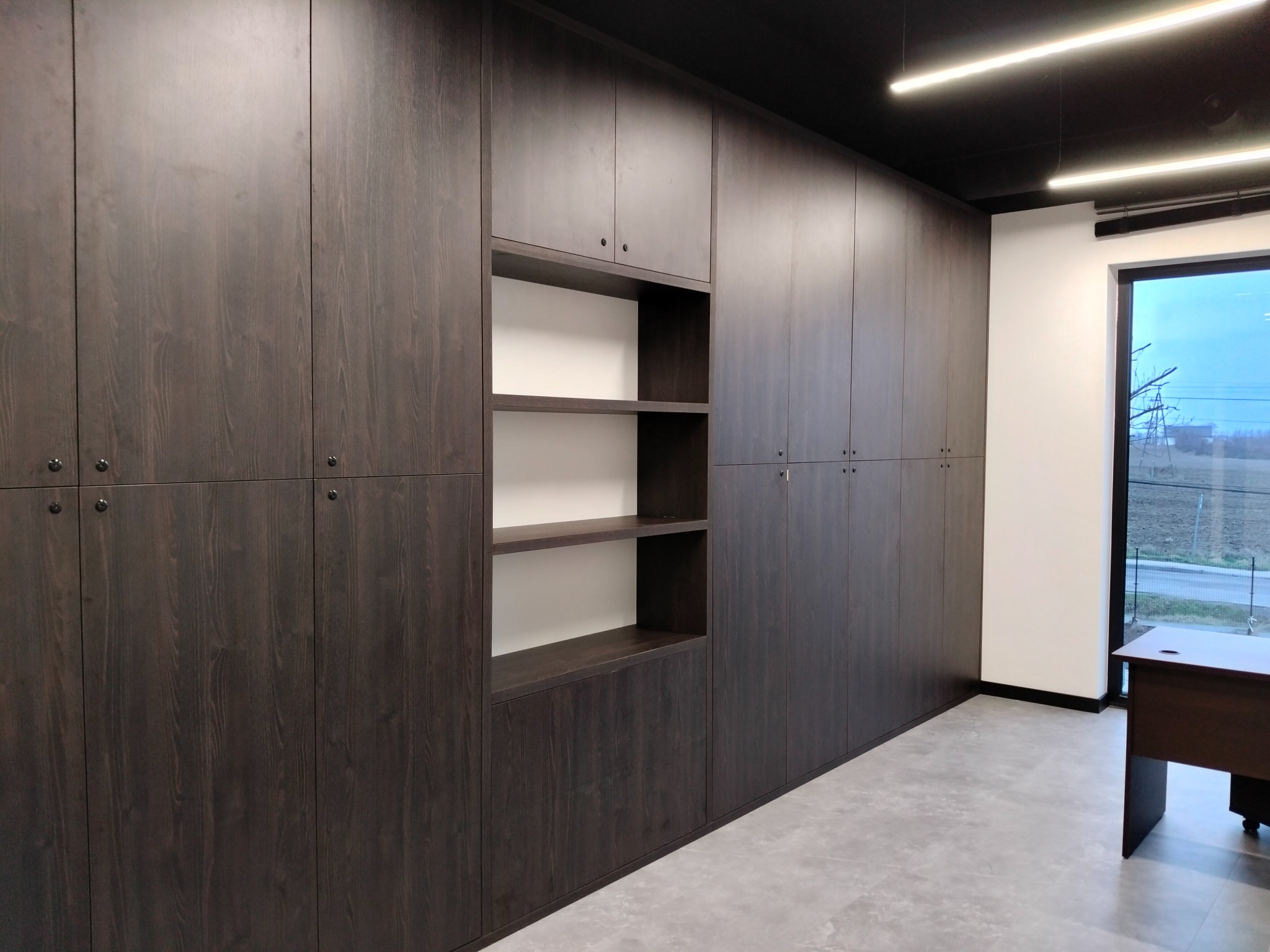 Długa ściana zabudowana ciemnymi szafami z drewnianymi frontami, z otwartą wnęką z półkami, oświetlenie sufitowe, szara podłoga