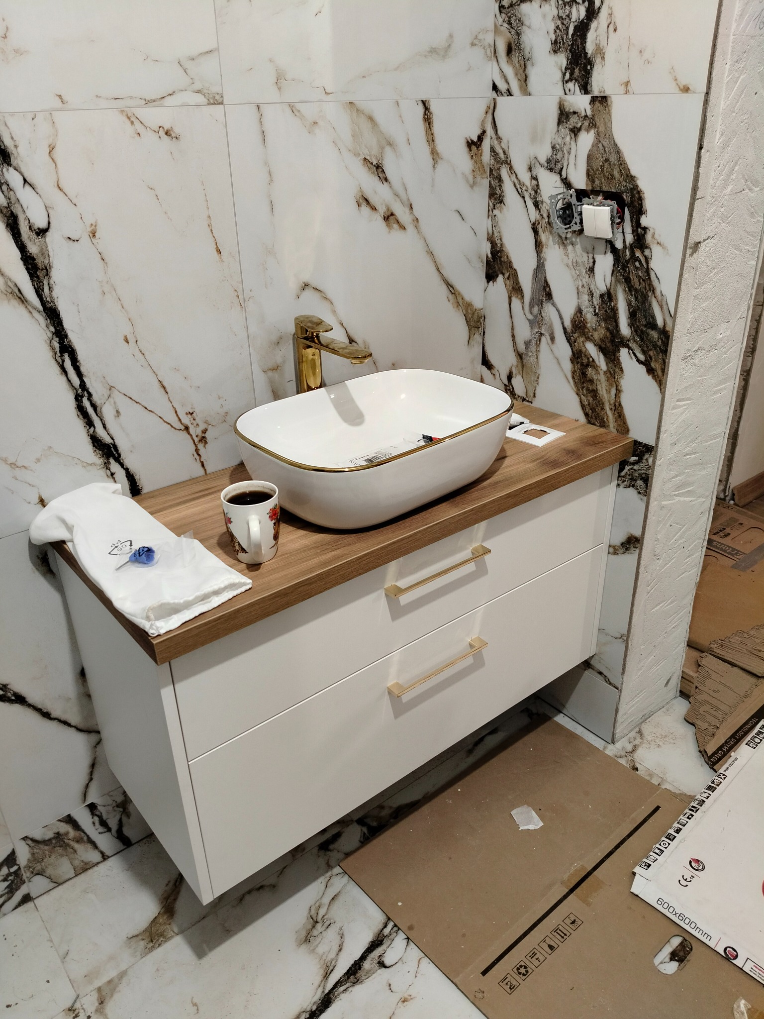 Biała umywalka nablatowa z złotym wykończeniem i baterią, zamontowana na białej szafce z drewnianym blatem, obok filiżanka kawy i ręcznik. W tle marmurowe płytki i widoczne prace wykończeniowe.
