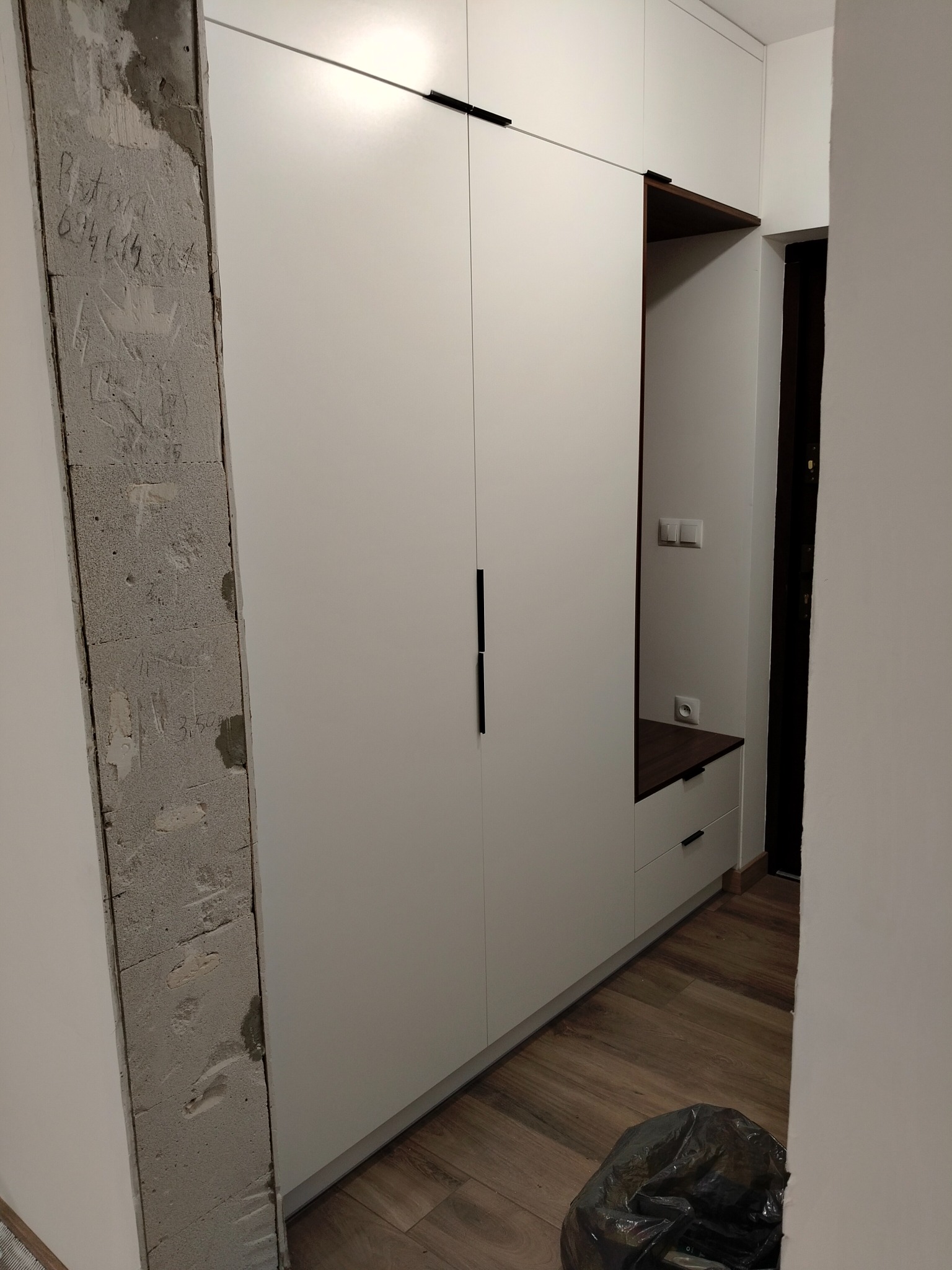 Biała szafa wnękowa z czarnymi uchwytami, widoczny fragment ściany z surowego betonu z napisami, drewnopodobna podłoga i czarny worek na śmieci w rogu pomieszczenia.