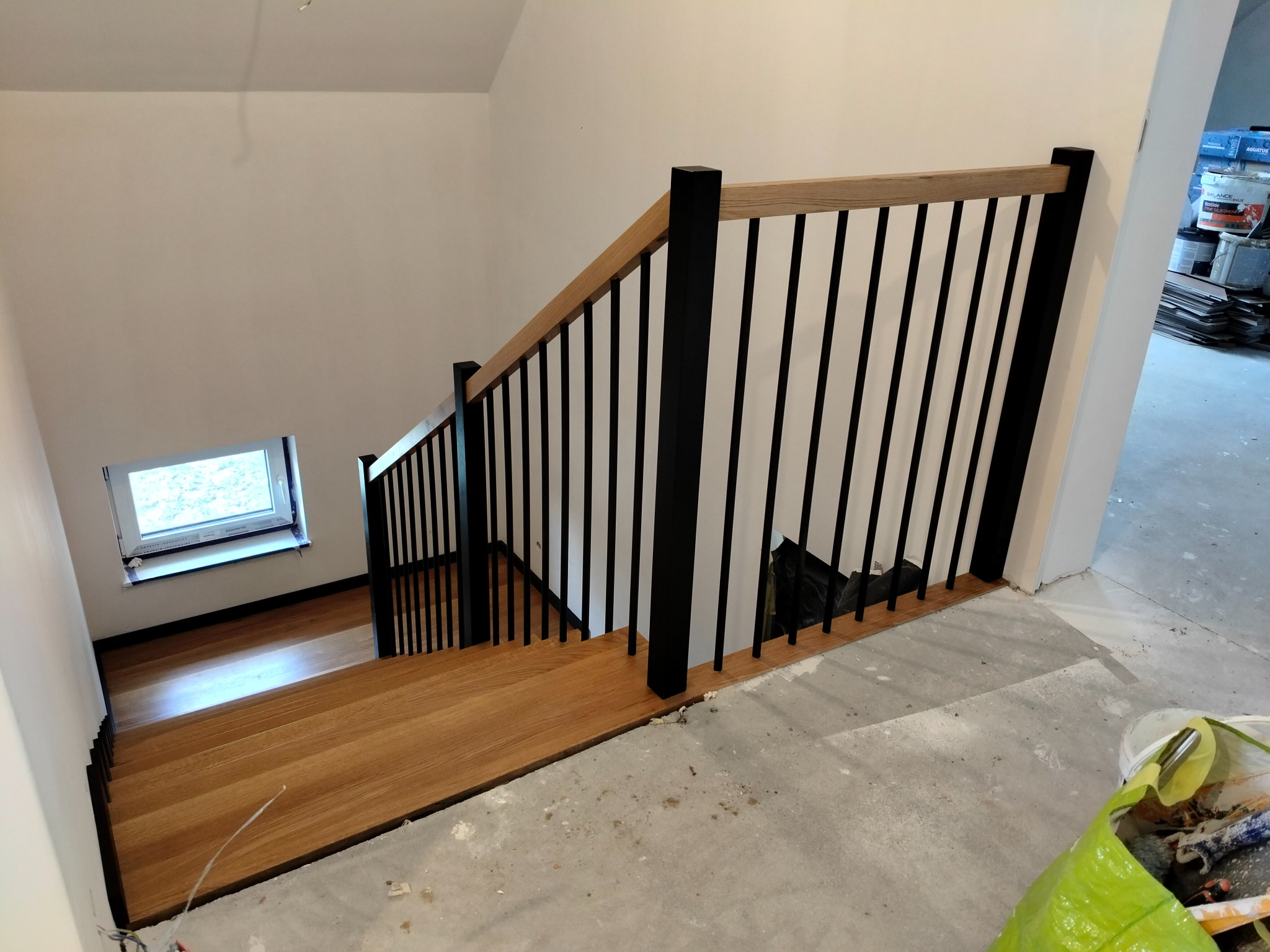 Nowoczesne schody z drewnianymi stopniami i czarną balustradą, widok z góry na dół, z oknem w tle i śladami prac wykończeniowych na podłodze.