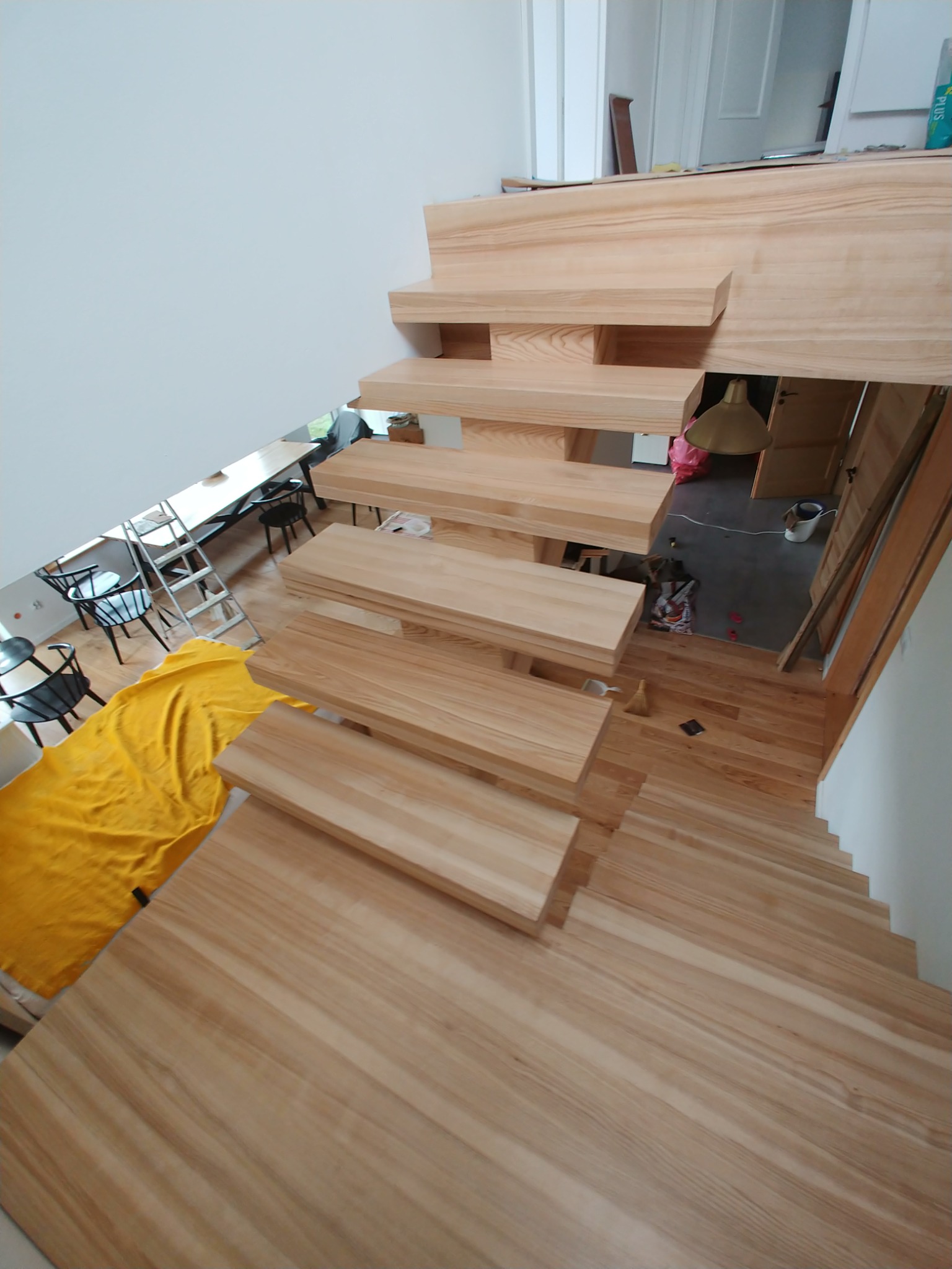 Szeroki kadr z góry na surowe drewniane schody w trakcie montażu, z widocznym zabezpieczeniem podłogi żółtą folią malarską i drabiną w tle.