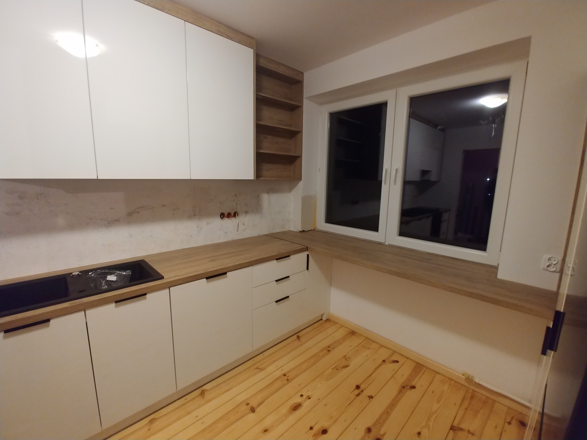 Białe szafki kuchenne z drewnianym blatem i otwartymi półkami, widoczny zlew i drewniana podłoga, ściana w trakcie wykończenia.