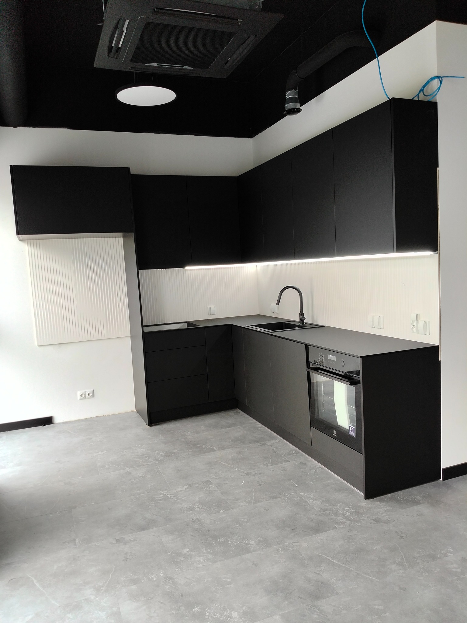 Nowoczesna, minimalistyczna kuchnia w czerni z szarą podłogą; widoczne czarne szafki kuchenne, czarny zlew z baterią, piekarnik oraz biała ściana z dekoracyjną fakturą.