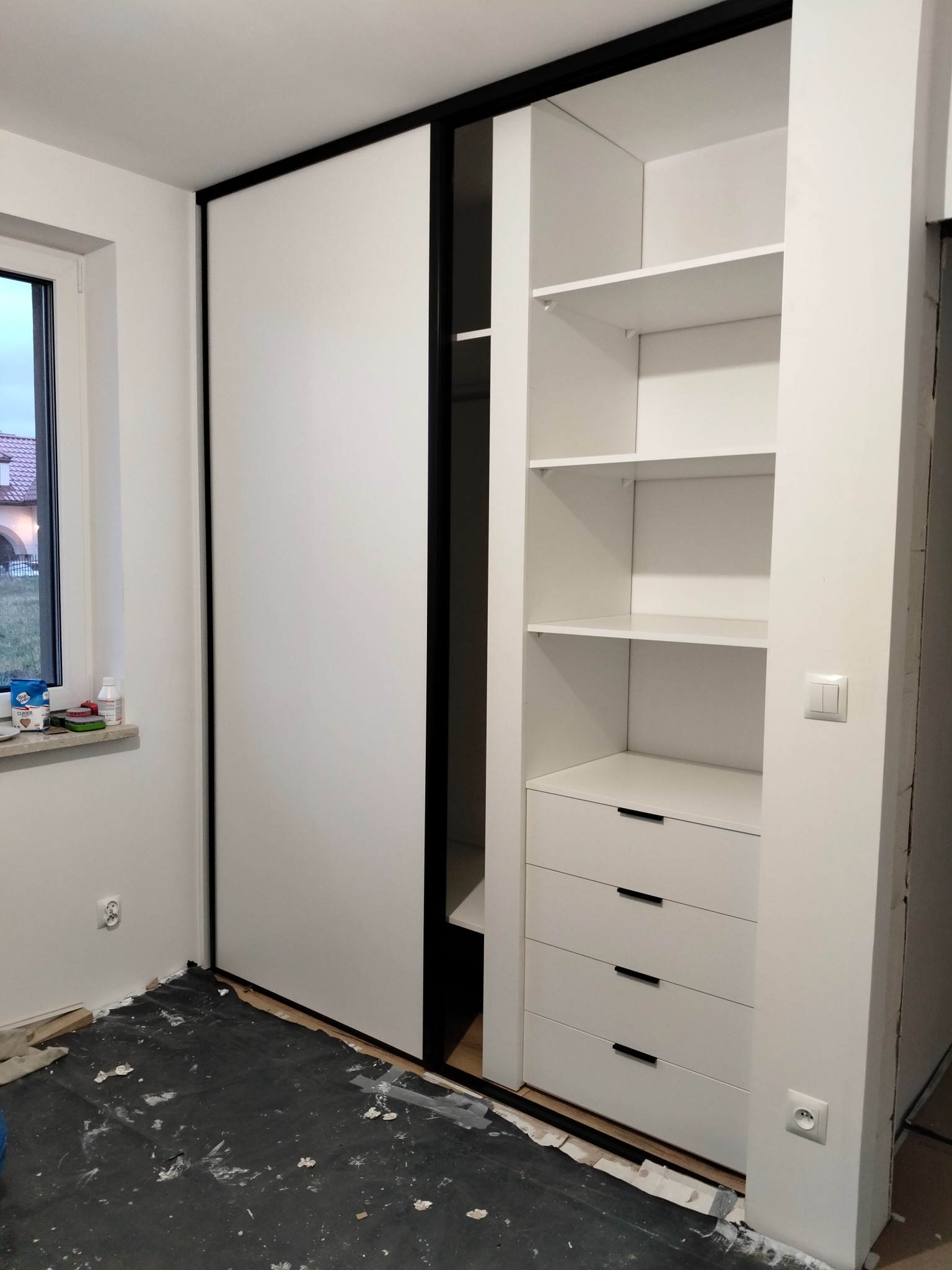 Biała szafa wnękowa z przesuwnymi drzwiami i czarnymi obramowaniami, widoczne półki i szuflady, podłoga zabezpieczona folią malarską, okno z widokiem na dom w tle.
