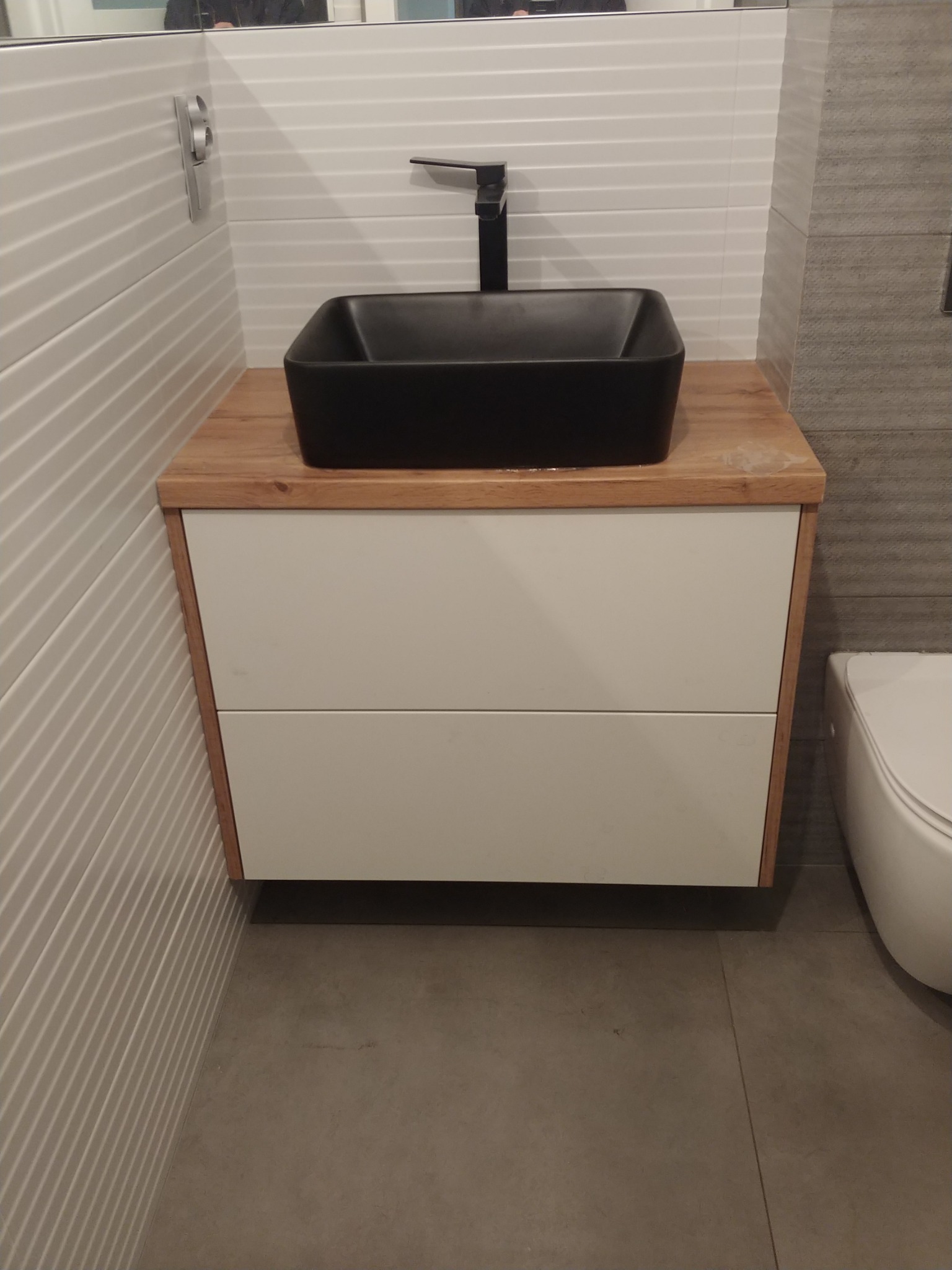 Łazienka z umywalką nablatową w kolorze czarnym, osadzoną na szafce z białymi frontami i drewnianym blatem. Widoczne białe, poziome płytki na ścianie i szara podłoga.