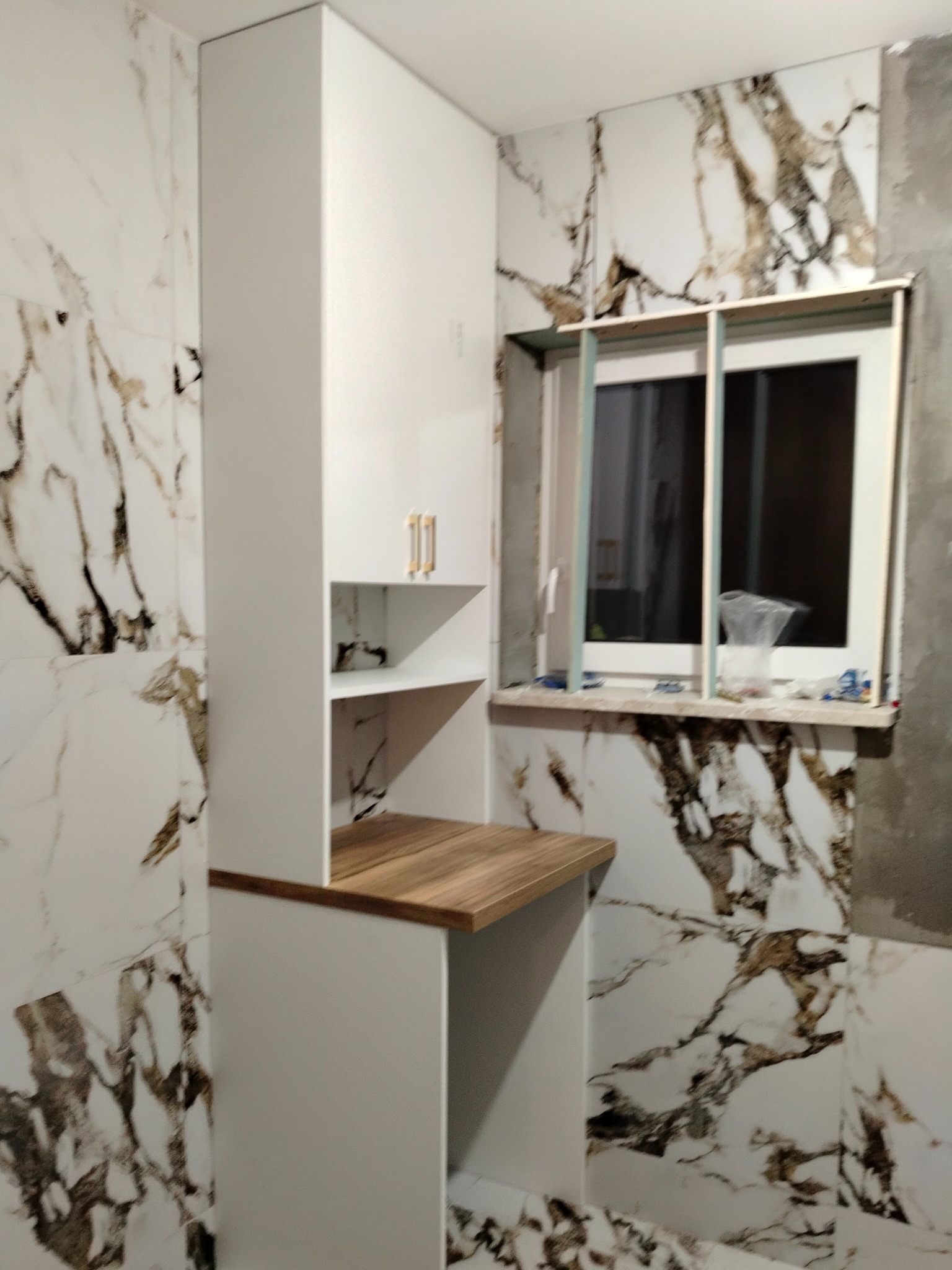 Biała szafka łazienkowa z drewnianym blatem, zamontowana przy oknie, na tle marmurowych płytek ściennych podczas remontu.