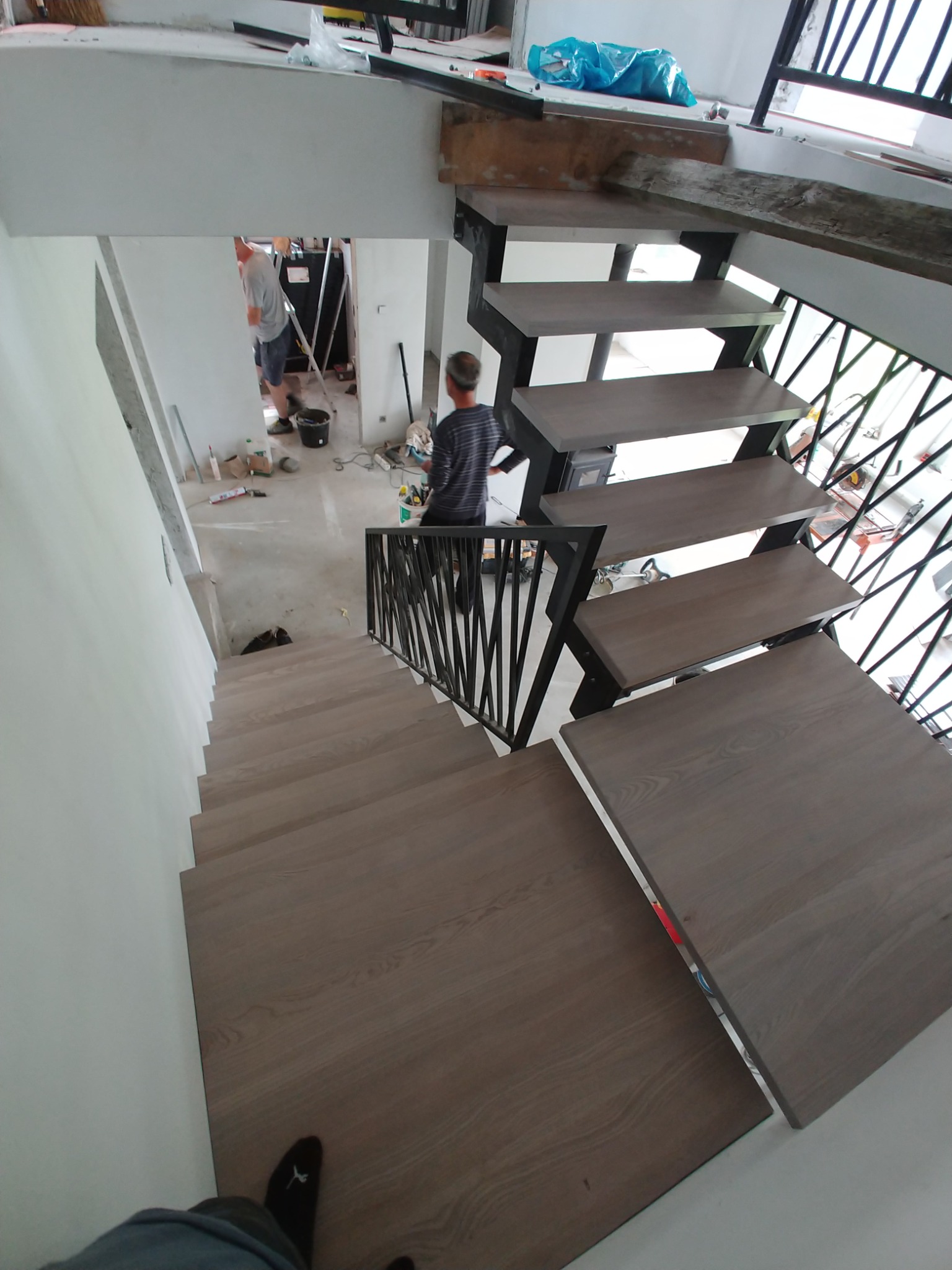Wnętrze w trakcie wykańczania: widok z góry na schody z drewnianymi stopniami i czarną, metalową balustradą o geometrycznym wzorze, w tle widoczni dwaj pracownicy podczas prac malarskich.
