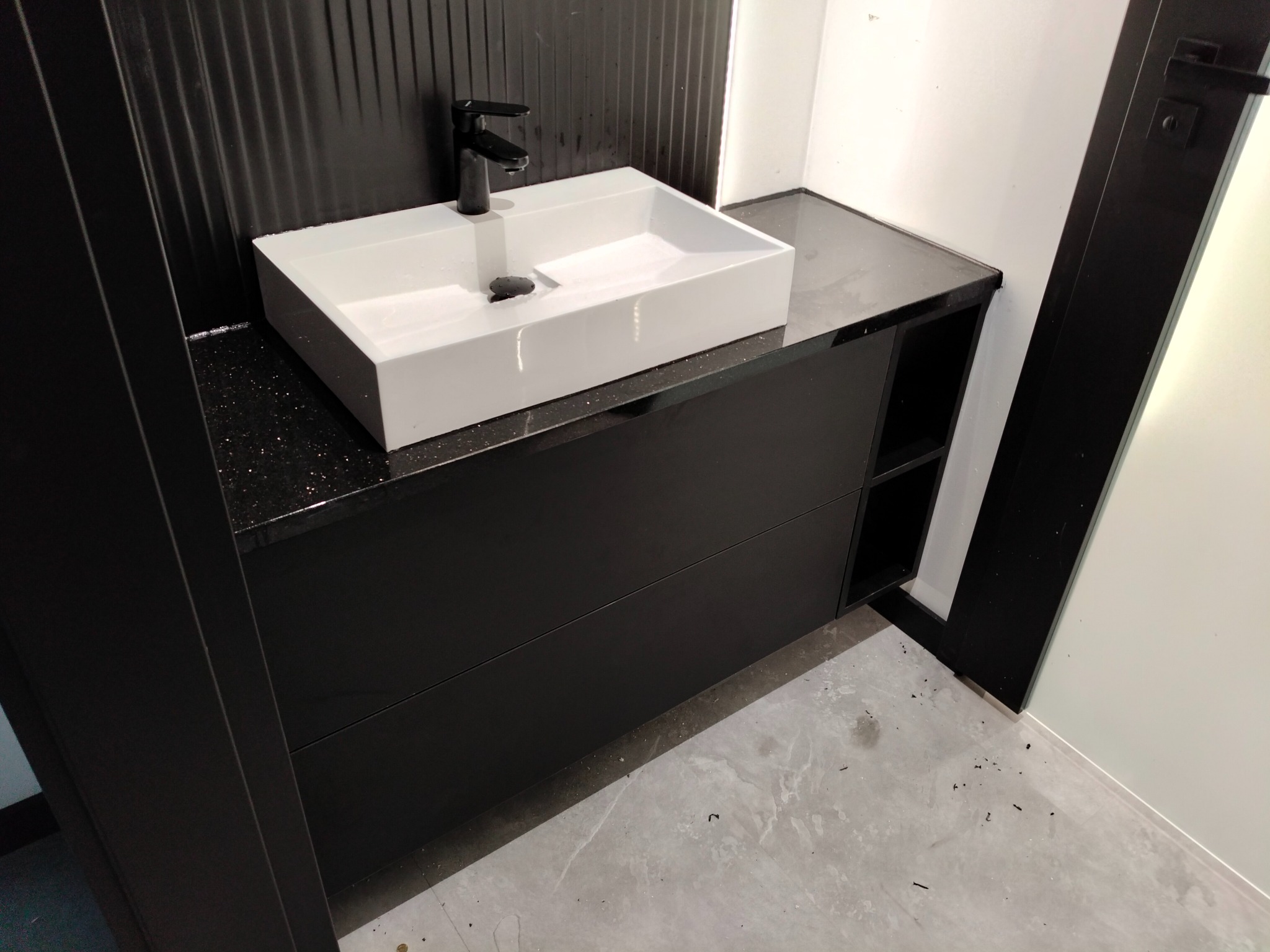 Nowoczesna, prostokątna umywalka nablatowa z czarną armaturą na czarnej szafce z blatem w czarne plamki, na tle czarnej ściany z pionowymi żłobieniami.