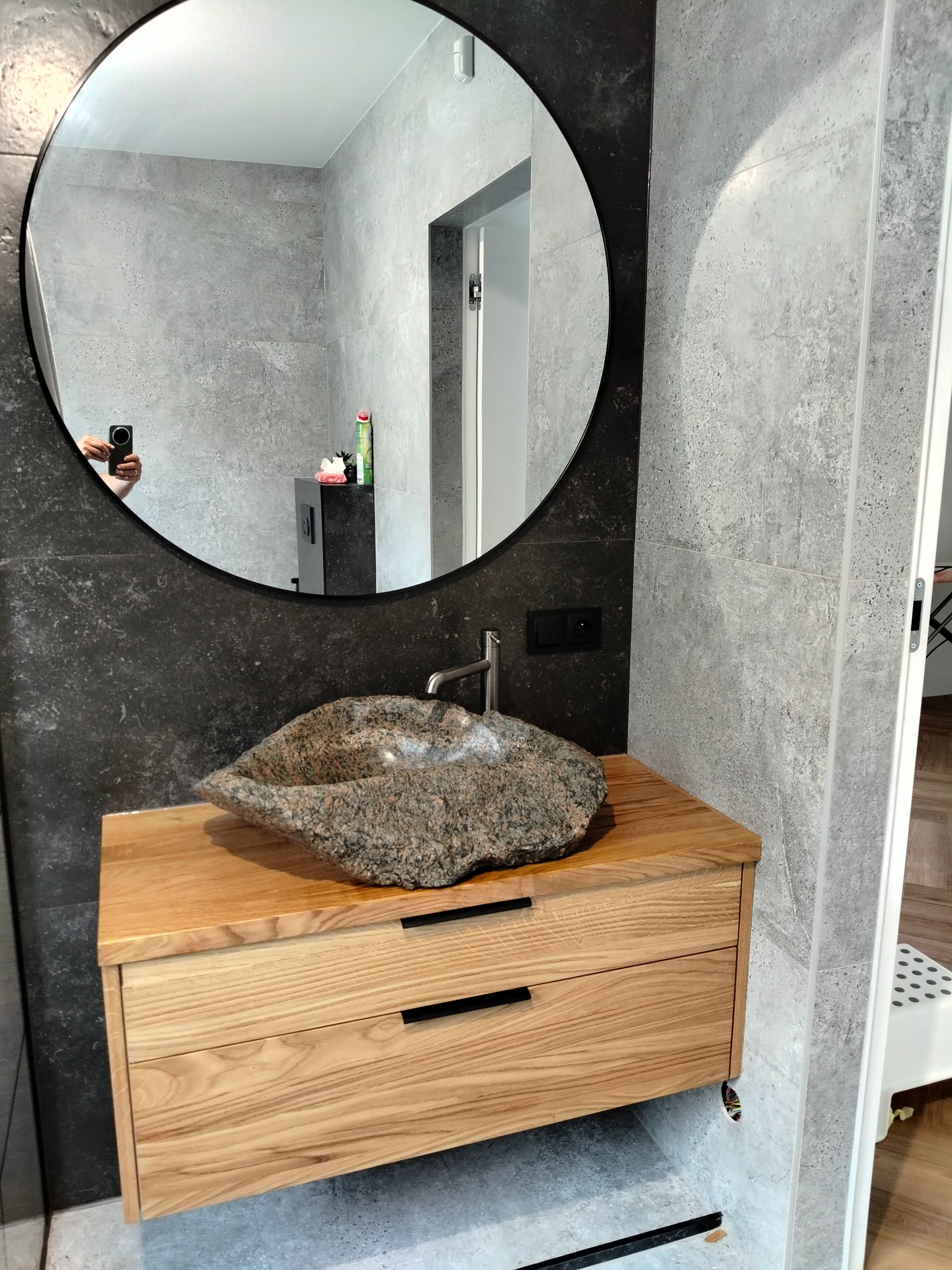 Łazienka z umywalką z kamienia na drewnianej szafce z dwiema szufladami, okrągłe lustro z czarną ramą odbija wnętrze pomieszczenia.