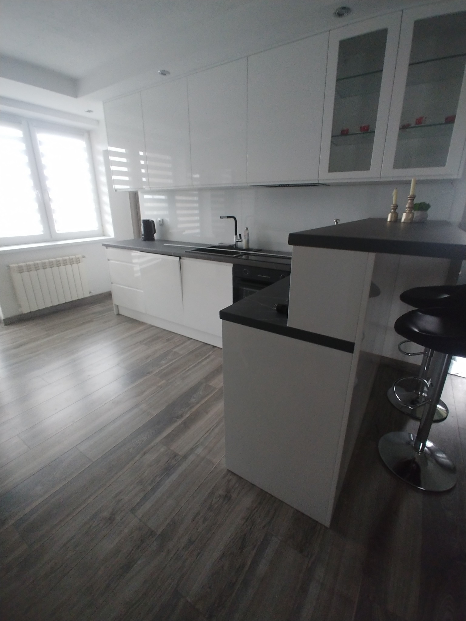 Biała, nowoczesna kuchnia z barkiem i hokerami. Fronty szafek na wysoki połysk, szare panele podłogowe imitujące drewno. Minimalistyczny design, jasne wnętrze.