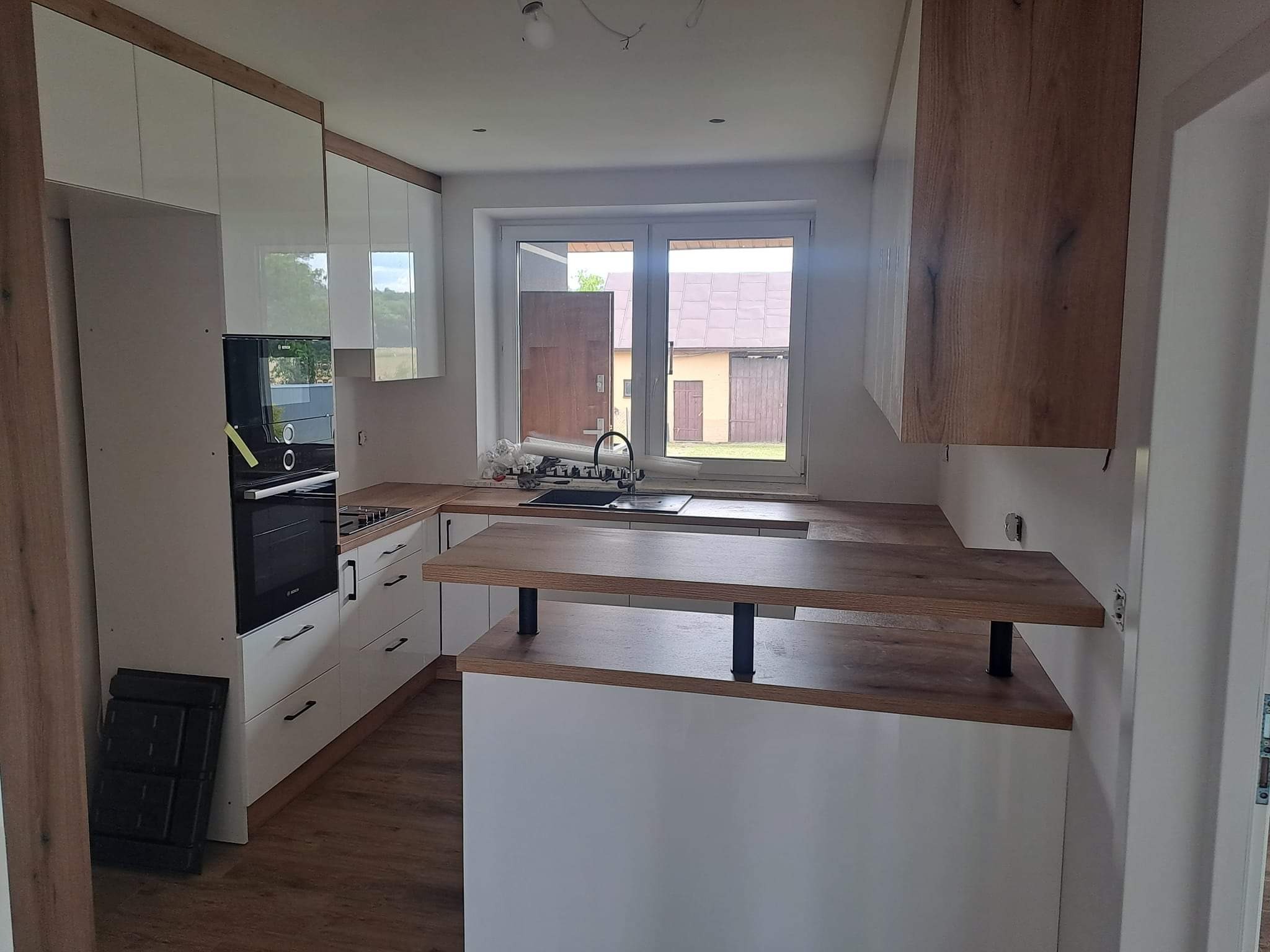 Jasna kuchnia z drewnianymi akcentami: białe szafki, drewniany blat i wyspa kuchenna z czarnymi wspornikami. Okno z widokiem na budynek gospodarczy.