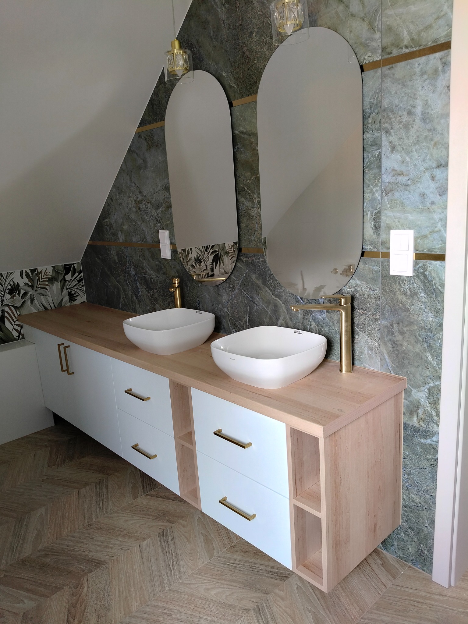 Nowoczesna łazienka na poddaszu z dwoma umywalkami nablatowymi, złotymi bateriami i owalnymi lustrami. Białe szafki z drewnianym blatem i złotymi uchwytami.