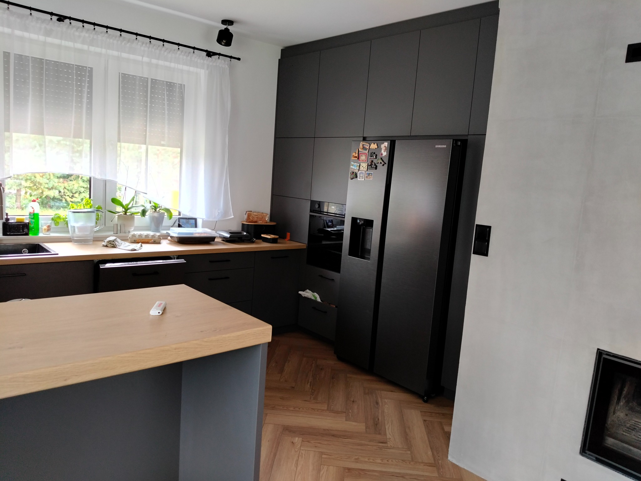 Nowoczesna kuchnia z szarymi szafkami, drewnianym blatem wyspy i parkietem w jodełkę. Widoczny czarny sprzęt AGD i okno z roślinami.