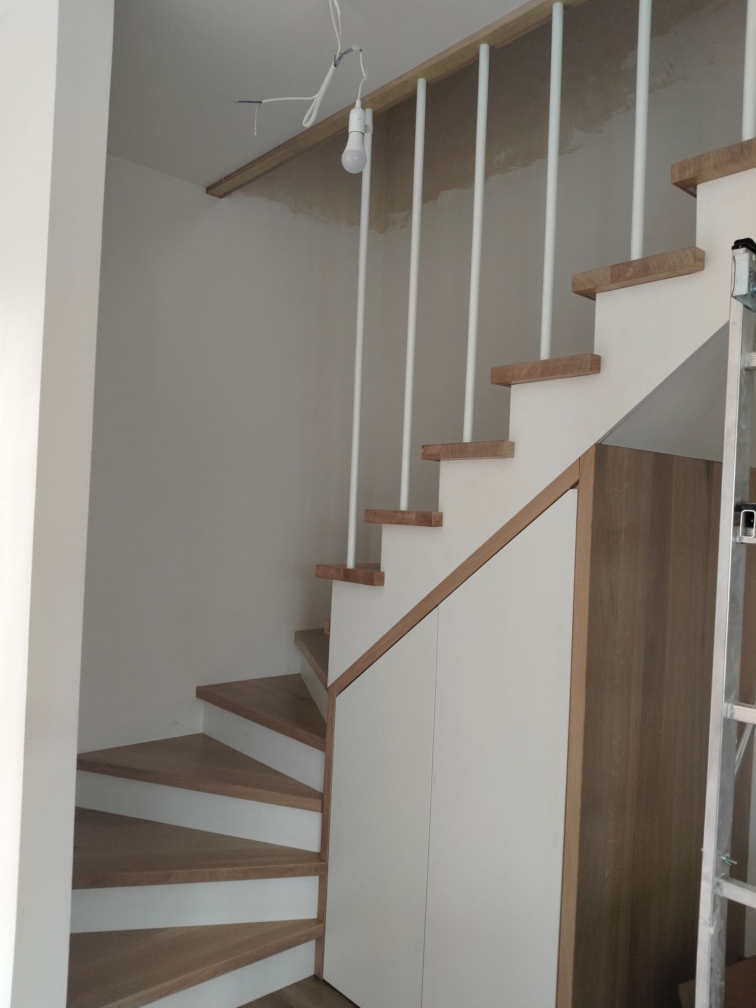 Drewniane schody spiralne z białą balustradą i zabudową szafy pod schodami. Widoczna drabina oparta o ścianę. Surowy stan wykończenia sufitu.