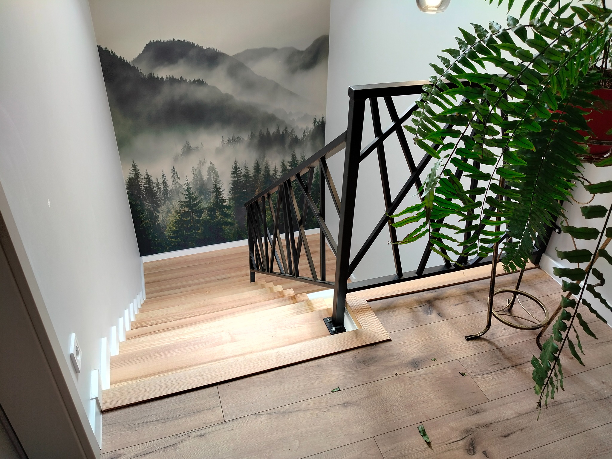 Nowoczesne schody drewniane z czarną balustradą w Krzeszowie. Na ścianie fototapeta z motywem górskim. U dołu widoczna podłoga z paneli i roślina doniczkowa.