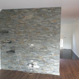 ŁUKASZ CYZ Budo - Solid - Ściana działowa wyłożona szarą okładziną kamienną w salonie, z widocznymi gniazdkami elektrycznymi i półkami z kamienia, podłoga z paneli imitujących drewno.