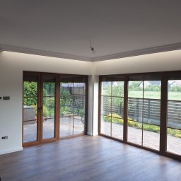 ŁUKASZ CYZ Budo - Solid - Przestronne wnętrze z dużymi oknami tarasowymi z drewnianą ramą, parkiet na podłodze, fragment ściany wykończony kamieniem dekoracyjnym, widoczna listwa sufitowa z oświetleniem LED i goła żarówka