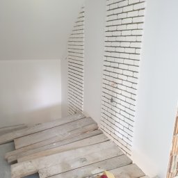 ŁUKASZ CYZ Budo - Solid - Ściana z białej cegły klinkierowej w trakcie montażu, widoczne drewniane deski zabezpieczające podłogę i akcesoria glazurnicze, surowy stan poddasza.